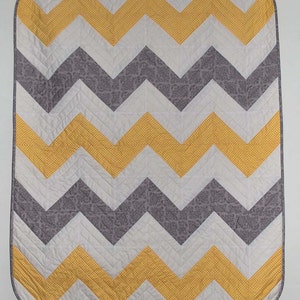 Pode incluir: Uma colcha com padrão chevron amarelo, cinza e branco. A colcha é feita de tecido de algodão e tem um design acolchoado.