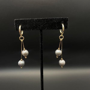 May include: Gold-coloured hoop earrings with a dangling design. Each earring has a small, round hoop connected to a delicate gold wire. Two grey, spherical beads are suspended from each wire, creating a layered effect. The earrings are displayed on a black stand.