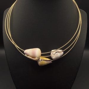 Könnte beinhalten: Eine goldfarbene Halskette mit drei Strängen, verziert mit drei Muschel-Anhängern in Rosa-, Weiß- und Gelbtönen. Die Halskette wird auf einer schwarzen Schaufensterpuppe präsentiert, die das Design und die Farbe des Schmucks hervorhebt.