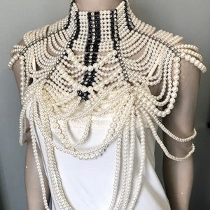 May include: An elaborate pearl and rhinestone shoulder piece. The design features multiple strands of off-white pearls cascading over the shoulders and chest. A high collar is accented with black and white stripes and rhinestones. The piece is displayed on a mannequin.