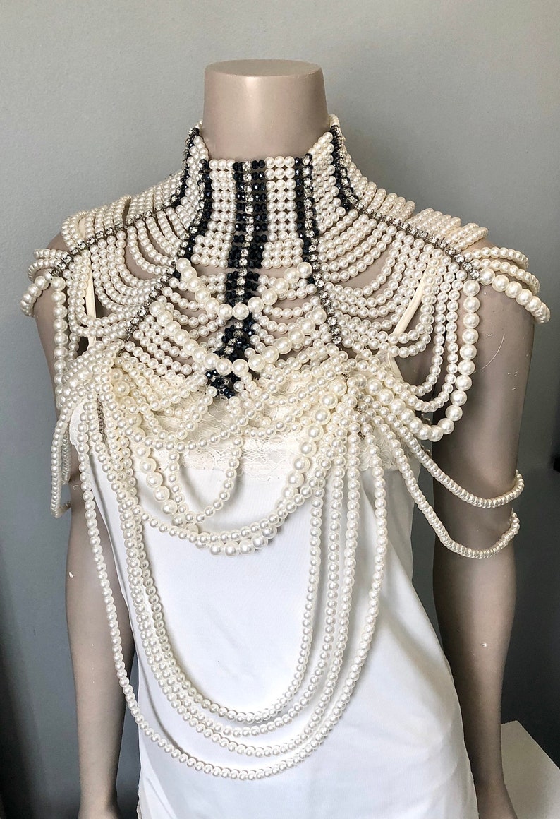 May include: An elaborate pearl and rhinestone shoulder piece. The design features multiple strands of off-white pearls cascading over the shoulders and chest. A high collar is accented with black and white stripes and rhinestones. The piece is displayed on a mannequin.