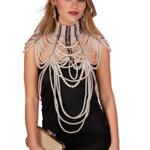 May include: A woman wearing a pearl necklace and earrings, paired with a black dress and a gold clutch. The necklace is a high-neck design with multiple strands of pearls cascading down the shoulders and chest. The clutch is rectangular with a beaded design.