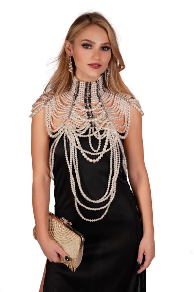 May include: A woman wearing a pearl necklace and earrings, paired with a black dress and a gold clutch. The necklace is a high-neck design with multiple strands of pearls cascading down the shoulders and chest. The clutch is rectangular with a beaded design.