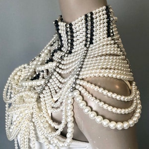 May include: A white pearl shoulder piece with a high collar. The collar features black and silver accents. Multiple strands of white pearls cascade down the shoulders. The piece is displayed on a mannequin.