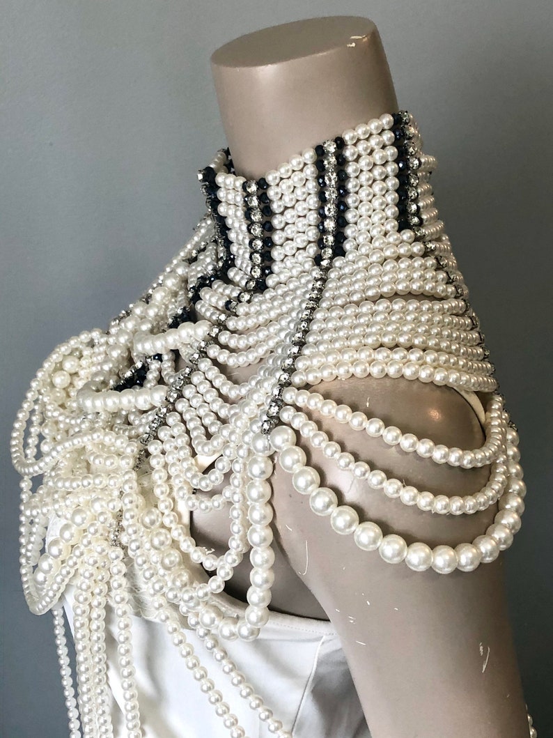 May include: A white pearl shoulder piece with a high collar. The collar features black and silver accents. Multiple strands of white pearls cascade down the shoulders. The piece is displayed on a mannequin.