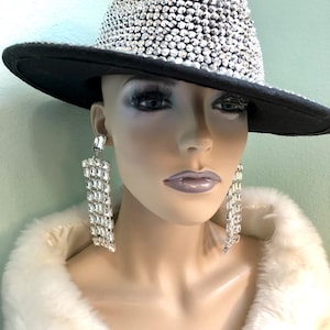 May include: A mannequin head wearing a black fedora hat covered in sparkling rhinestones. The hat has a black brim. The mannequin is also wearing long, dangling rhinestone earrings and a white faux fur wrap.