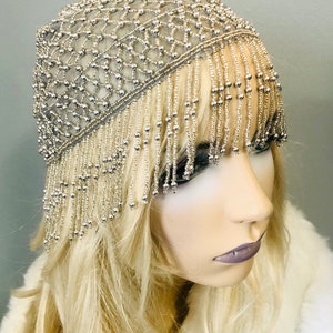 May include: Silver beaded headpiece with a net design and dangling fringe. The headpiece is displayed on a mannequin with blonde hair and a white fur-like garment. The headpiece is a vintage style.