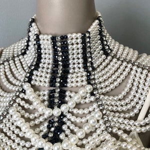 May include: A white and black beaded collar necklace. The necklace features multiple strands of white faux pearls, interspersed with black beads and clear rhinestones. The collar is displayed on a mannequin.