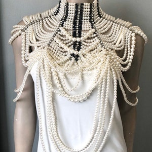 May include: An elaborate pearl and rhinestone shoulder piece. The design features multiple strands of off-white pearls cascading from a high, structured collar with black and white striped accents. Rhinestones add sparkle.