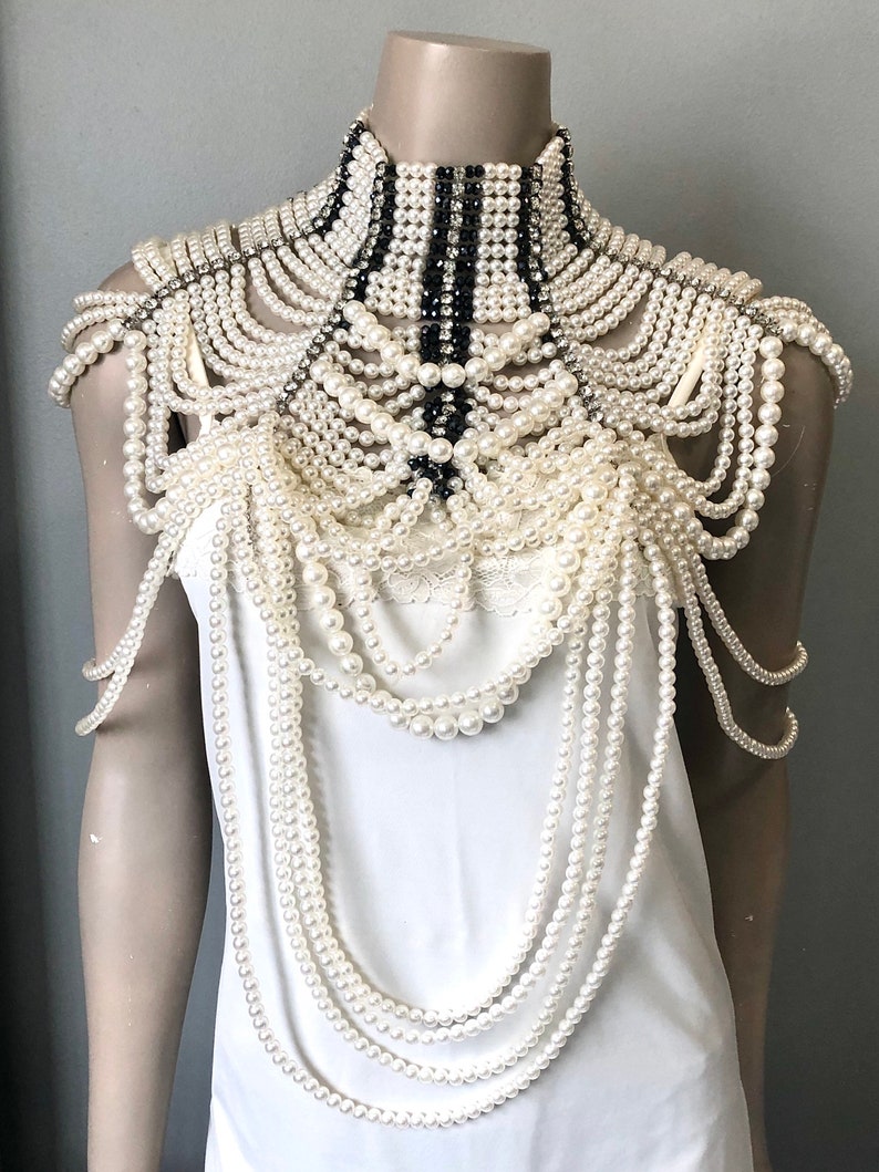 May include: An elaborate pearl and rhinestone shoulder piece. The design features multiple strands of off-white pearls cascading from a high, structured collar with black and white striped accents. Rhinestones add sparkle.