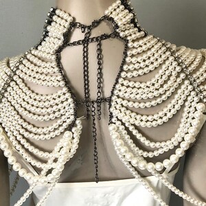 May include: A white pearl and chain shoulder piece. The piece features multiple strands of white pearls cascading down the shoulders, connected by dark metal chains. The collar is lined with small crystals and pearls.