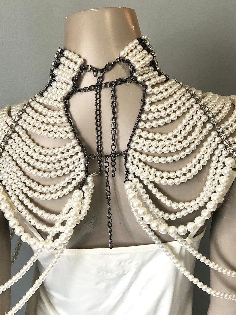 May include: A white pearl and chain shoulder piece. The piece features multiple strands of white pearls cascading down the shoulders, connected by dark metal chains. The collar is lined with small crystals and pearls.