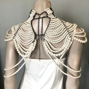 May include: A pearl shoulder necklace with multiple strands of white faux pearls cascading over the shoulders. The necklace is attached with dark metal chains and accents, creating a dramatic and elegant look. The necklace is displayed on a mannequin.