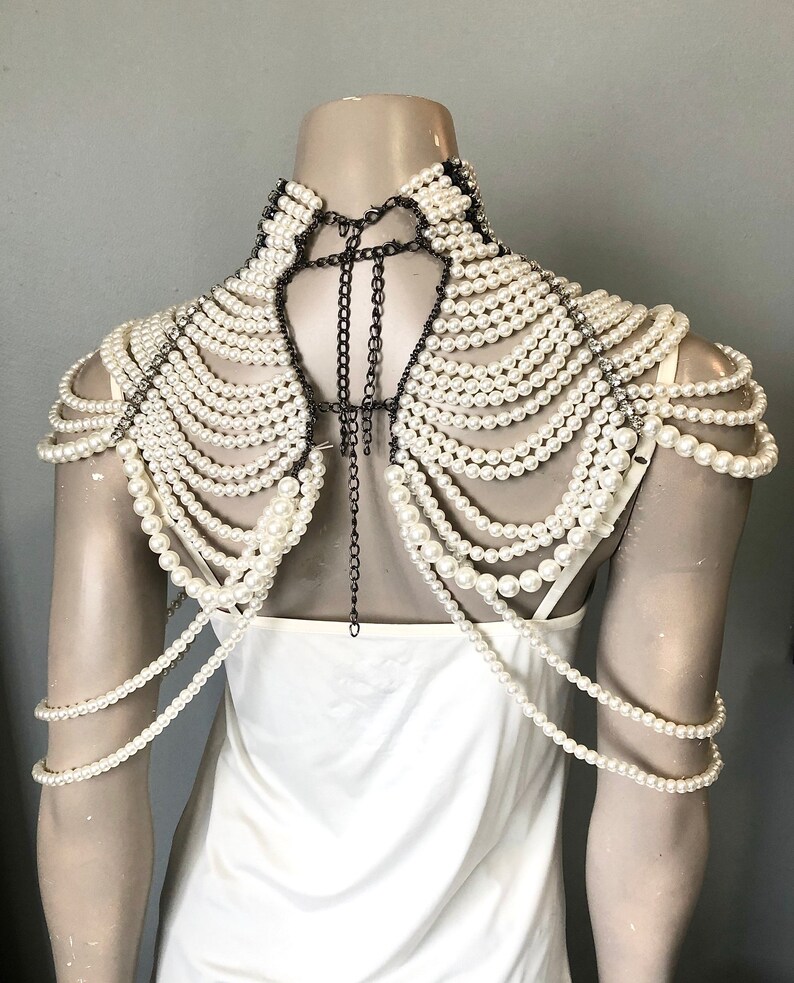May include: A pearl shoulder necklace with multiple strands of white faux pearls cascading over the shoulders. The necklace is attached with dark metal chains and accents, creating a dramatic and elegant look. The necklace is displayed on a mannequin.