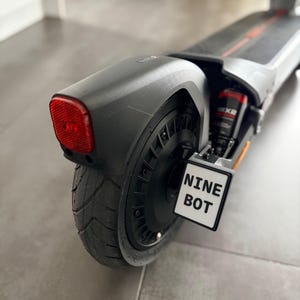 Könnte beinhalten: Schwarzer und grauer Elektroroller mit rotem Rücklicht und dem Text "NINE BOT" auf einem weißen Schild. Der Roller hat einen schwarzen Reifen und ein Federungssystem.
