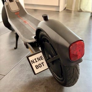 Op de afbeelding: Een grijze elektrische step met een zwart achterspatbord, een rode reflector en een witte rechthoekige kentekenplaathouder met de zwarte tekst NINE BOT bevestigd aan het achterwiel. De step heeft een gestructureerd voetdek met rode accenten.
