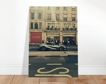 May include: A photograph of a silver sports car driving on a city street. The car is in front of a building with multiple stories and windows. The image is displayed on a wooden floor.