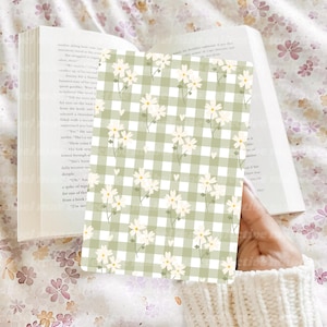 Puede incluir: Un cuaderno con estampado floral con un fondo de cuadros verdes y flores de margarita blancas. El cuaderno se sostiene abierto frente a un libro. El fondo tiene un estampado floral.