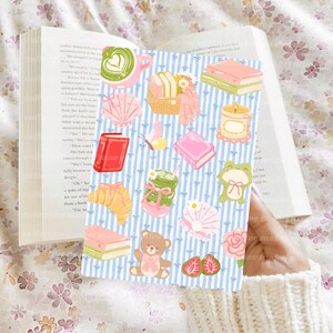 May include: A rectangular card featuring a light blue and white vertical striped background with various illustrations including a green latte, a picnic basket, books, a candle, a frog, a teddy bear, seashells, a croissant, and strawberries.