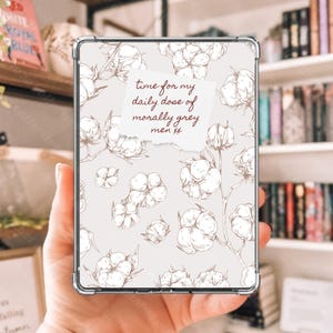 May include: A clear phone case with a floral design featuring cotton branches and the text "time for my daily dose of morally grey men xx" on a light beige background. The case is held in a hand.