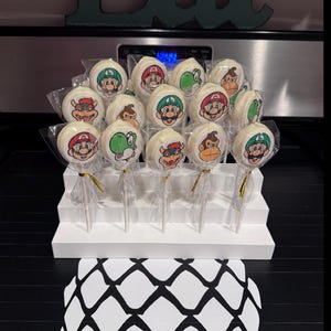 May include: A collection of white cake pops featuring various cartoon character designs, including Mario, Luigi, Yoshi, and Donkey Kong. The pops are arranged on a white tiered display, with gold ribbon accents. The background includes a black surface and a teal-colored word art.