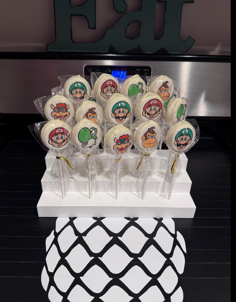 May include: A collection of white cake pops featuring various cartoon character designs, including Mario, Luigi, Yoshi, and Donkey Kong. The pops are arranged on a white tiered display, with gold ribbon accents. The background includes a black surface and a teal-colored word art.