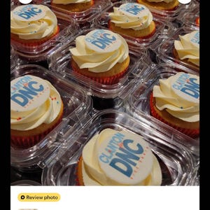 May include: Cupcakes with white frosting and edible toppers featuring the text "Clanton DNC" in blue and white. The cupcakes are in clear plastic containers, arranged in rows. The frosting is swirled, and the cupcake liners are red.