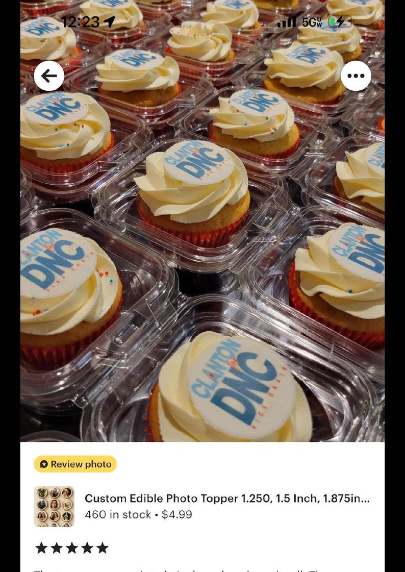 May include: Cupcakes with white frosting and edible toppers featuring the text "Clanton DNC" in blue and white. The cupcakes are in clear plastic containers, arranged in rows. The frosting is swirled, and the cupcake liners are red.