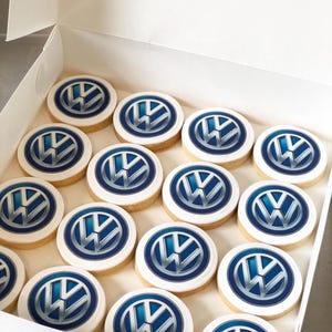 May include: A box of round cookies with the Volkswagen logo. Each cookie has a white icing border and a blue and silver logo in the center. The cookies are arranged neatly inside a white box.
