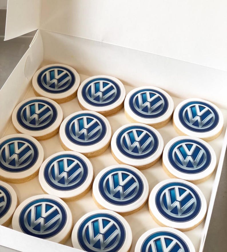 May include: A box of round cookies with the Volkswagen logo. Each cookie has a white icing border and a blue and silver logo in the center. The cookies are arranged neatly inside a white box.