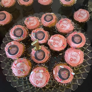 May include: A collection of cupcakes with pink frosting, each topped with a photo and the words "Sweet One." Some cupcakes have sprinkles and a gold number one candle. The cupcakes are arranged on a clear, decorative platter.