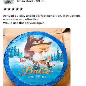May include: A round, edible cake topper featuring the animated film "Balto." The design includes the film's title, characters, and a blue border. The topper is placed on a brown surface.