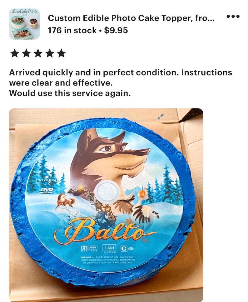 May include: A round, edible cake topper featuring the animated film "Balto." The design includes the film's title, characters, and a blue border. The topper is placed on a brown surface.