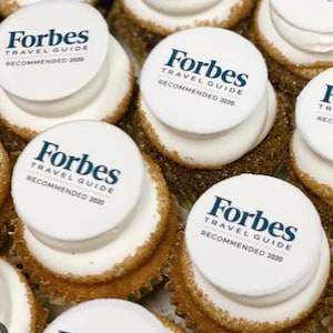 May include: Close-up of several cupcakes with white frosting and a sprinkle of brown sugar around the edges. Each cupcake is topped with a white circular edible image that reads "Forbes Travel Guide Recommended 2020" in dark blue text.