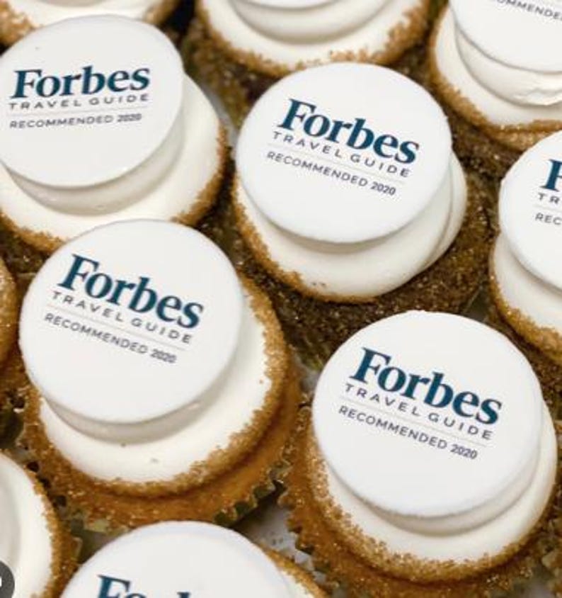 May include: Close-up of several cupcakes with white frosting and a sprinkle of brown sugar around the edges. Each cupcake is topped with a white circular edible image that reads "Forbes Travel Guide Recommended 2020" in dark blue text.