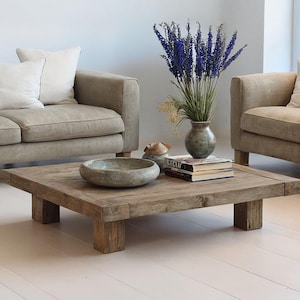 May include: Two beige sofas with white pillows face each other, flanking a square, rustic wooden coffee table. A vase of purple flowers and books sit on the table. A decorative bowl is also on the table. The room has white walls and flooring.