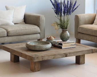 May include: Two beige sofas with white pillows face each other, flanking a square, rustic wooden coffee table. A vase of purple flowers and books sit on the table. A decorative bowl is also on the table. The room has white walls and flooring.