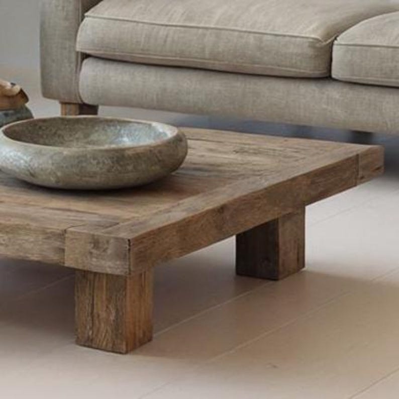 May include: A rustic, square coffee table made of weathered wood. The table has a thick, plank-style top and sturdy, block-style legs. A gray stone bowl sits on the table's surface. The table is near a light-colored sofa.
