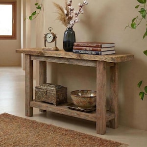 May include: A rustic wooden console table with a lower shelf, holding a decorative box and a patterned bowl. The top shelf displays a clock, books, and a vase with cotton stems. The table is made of weathered wood.
