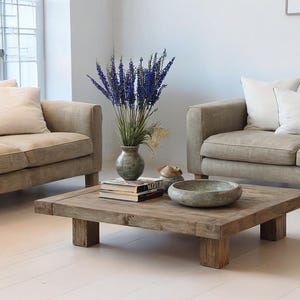May include: A living room scene featuring two beige sofas with cream-colored pillows. A square, rustic wooden coffee table holds books, a decorative bowl, and a vase of purple flowers. The room has white walls and light-colored flooring.