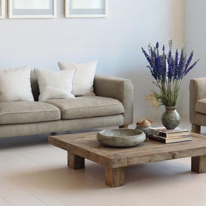 May include: A living room scene featuring two beige sofas with white pillows, a square wooden coffee table, and a vase of purple flowers. The room has a minimalist design with white walls and floors.