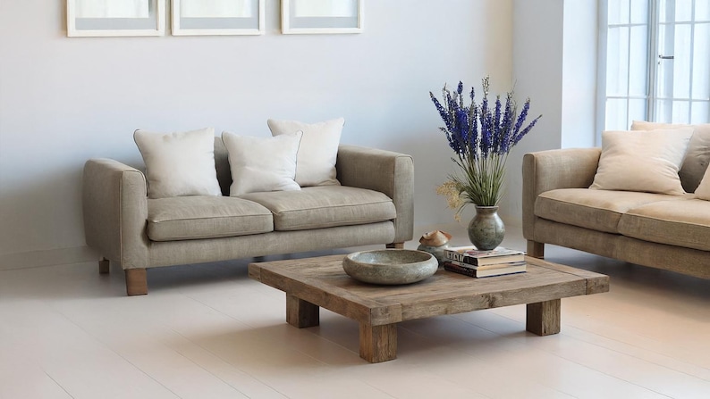 May include: A living room scene featuring two beige sofas with white pillows, a square wooden coffee table, and a vase of purple flowers. The room has a minimalist design with white walls and floors.