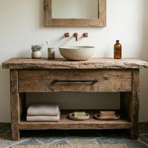 Può includere: Un mobile da bagno in legno rustico dotato di un cassetto con maniglia nera e un ripiano inferiore aperto. Il mobile sostiene un lavabo da appoggio beige maculato, una piccola pianta grassa, un portaspazzolino e una bottiglia in vetro marrone.