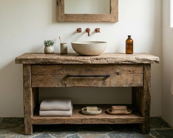 Puede incluir: Un mueble de baño de madera rústica con un cajón con tirador negro y un estante inferior abierto. El mueble sostiene un lavabo de sobreponer beige moteado, una pequeña planta suculenta, un portacepillos y una botella de vidrio marrón.