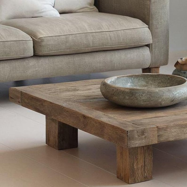 May include: A rustic, square coffee table made of weathered wood, featuring a thick tabletop and sturdy block legs. A stone bowl sits on the table, next to a beige sofa with a white pillow. The table's dimensions are approximately 40 inches square.
