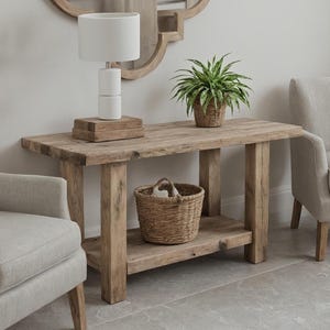 May include: A rustic wooden console table with a lower shelf, supporting a woven basket. A white lamp sits on top, along with a small plant in a woven pot. The table is made of light brown wood.