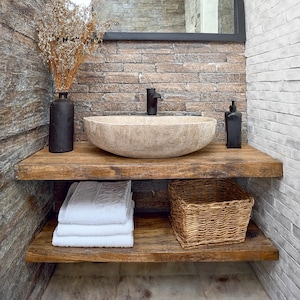 May include: A rustic bathroom vanity with a stone vessel sink, black faucet, and a black soap dispenser. The vanity has two wooden shelves with white towels and a woven basket. A black vase holds dried flowers. The background features brick and stone walls.