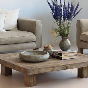 May include: A rustic, square wooden coffee table with a low profile, featuring a natural wood grain finish. The table is surrounded by beige sofas with white pillows, a stone bowl, a vase of flowers, and books.
