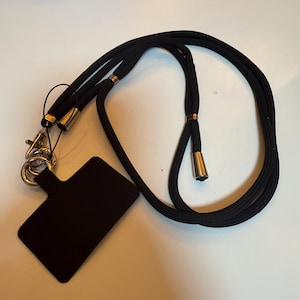 May include: Black phone lanyard with a black rectangular phone holder. The lanyard is made of a thick, woven black cord with gold-coloured metal accents and a metal clasp. The phone holder is a solid black colour.