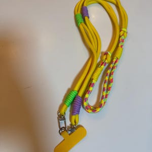 Puede incluir: Un cordón amarillo con un clip de plástico amarillo. El cordón está hecho de cuerda trenzada con detalles verdes, morados y rosas.